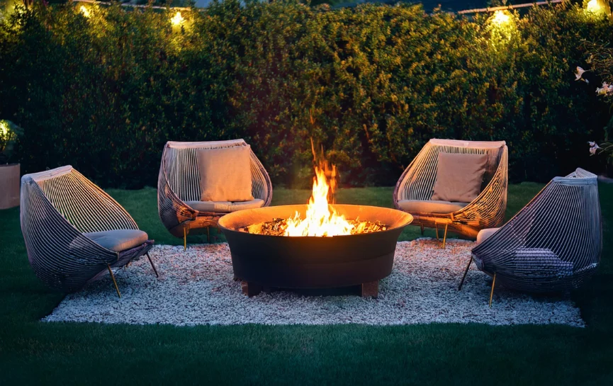 Muebles de exterior alrededor de una fogata de hierro para exterior encendida en un jardín iluminado por la noche.